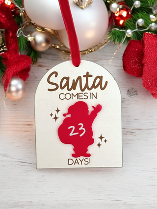 A red and white Santa Countdown Ornament with 'Santa comes in' and a number counting down written on it, hanging from a Christmas tree with holiday decorations around.